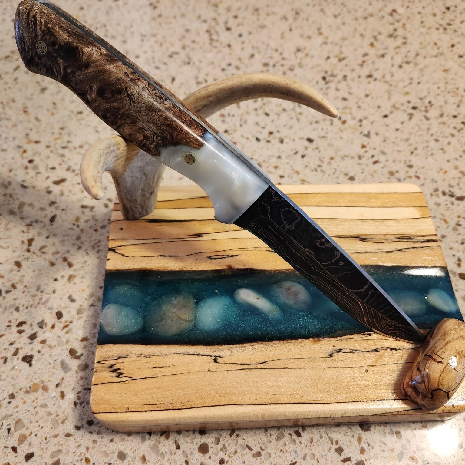 Decorative knife with wooden handle and blade on a wooden stand with blue resin design, on a speckled surface.