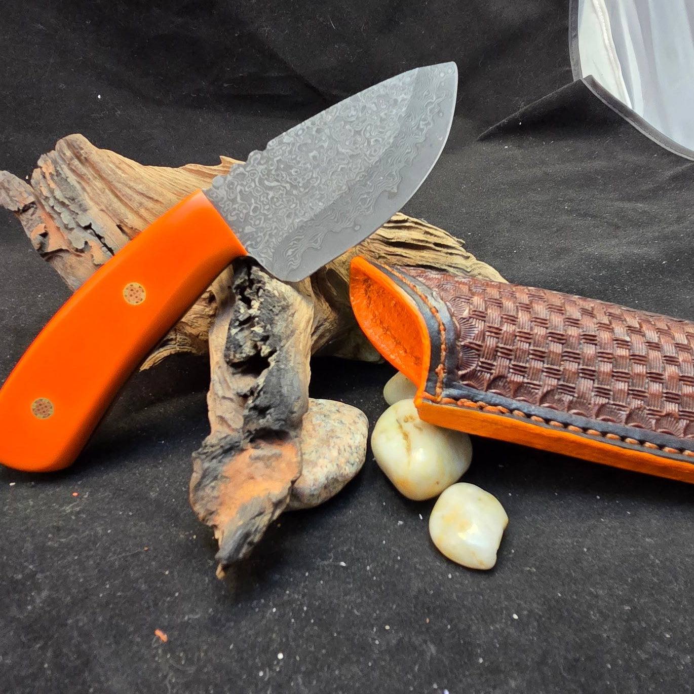 Orange-handled knife with a textured blade and leather sheath on a dark surface with driftwood and rocks