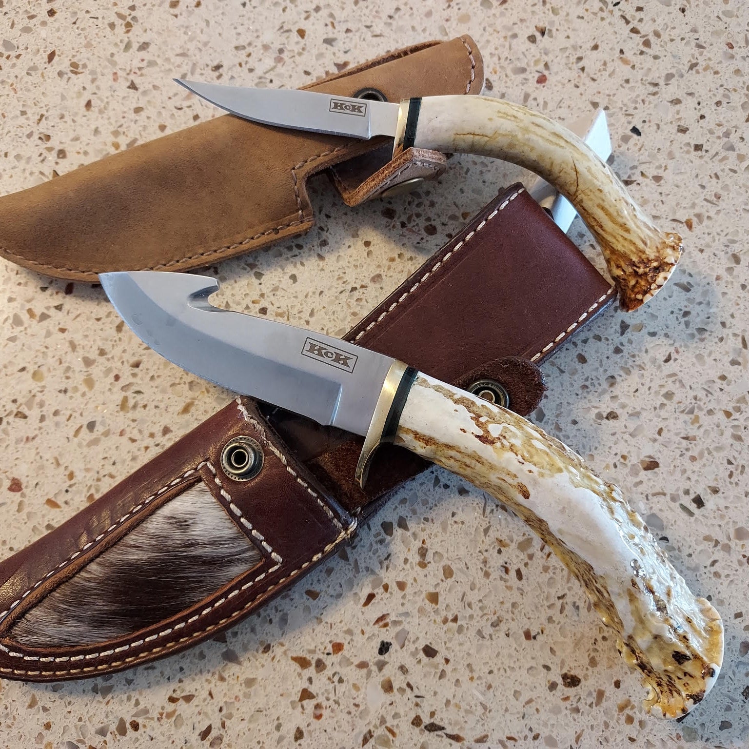 Two hunting knives made from deer antlers with leather sheaths on a speckled surface