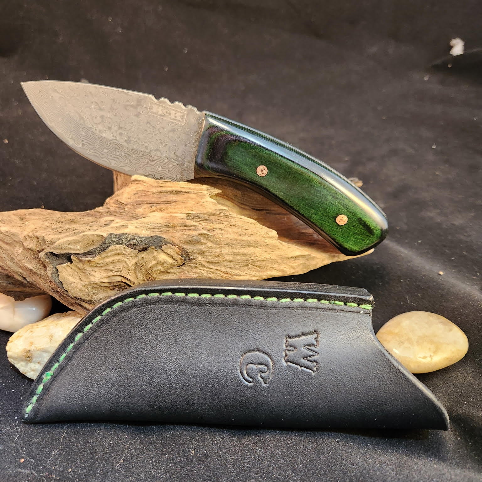 Knife with green handle and black sheath on a dark surface with wood and stones.